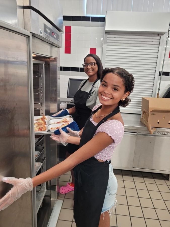 Mariah (24)(front) and Giselle (23)(back) seen helping out the cafeteria staff by crafting some lunches for the student body.